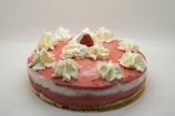 A round cake with a soft pink frosting layer and a middle layer of creamy white filling. The top is adorned with several dollops of white whipped cream and a single fresh strawberry placed in the center.