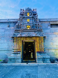 A detailed view of a traditional temple entrance adorned with intricate carvings and sculptures. The structure has a gopuram with multiple tiers, each with ornate details. Yellow and orange marigold garlands hang by the doorway, and the entrance is dimly lit, suggesting evening or nighttime. The temple is made of stone, and the sky is clear and blue.