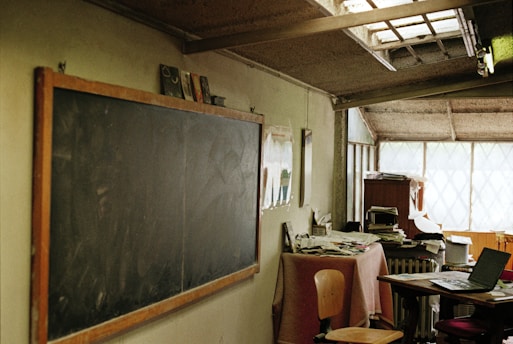 A classroom setting with an old wooden-framed blackboard on the left wall. A table covered in a pink cloth is cluttered with books and papers, accompanied by two chairs. In the background, a desk with more disorganized papers and a laptop is visible. The room has a rustic, dimly lit atmosphere with textured walls and a small window allowing limited natural light.