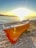 A small boat with Greek lettering is resting on a pebbly beach, bathed in the warm glow of a setting sun. The calm sea reflects the vivid colors of the sky, creating a peaceful and serene atmosphere.