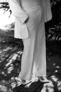 A person is wearing an elegant white suit and high-heeled shoes. The suit has a tailored jacket with relaxed fit pants, and the person has one hand in their pocket. They are standing on a paved surface with dappled sunlight creating patterns on the ground.