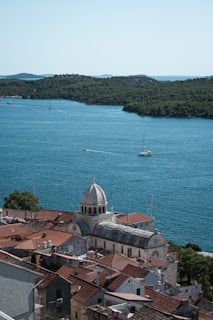 A scenic view of a coastal town with a prominent historical cathedral featuring a dome and red-tiled roofs. The town is set against a backdrop of calm blue waters and a lush green coastline. Several boats are visible on the water, emphasizing the tranquil and picturesque setting.