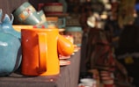 A variety of colorful pottery items, including mugs and vases, are displayed on a table. The focus is on a bright orange mug with a whimsical design, alongside a blue ceramic piece. The background is filled with more pottery, creating a vibrant market scene.