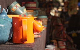 A variety of colorful pottery items, including mugs and vases, are displayed on a table. The focus is on a bright orange mug with a whimsical design, alongside a blue ceramic piece. The background is filled with more pottery, creating a vibrant market scene.
