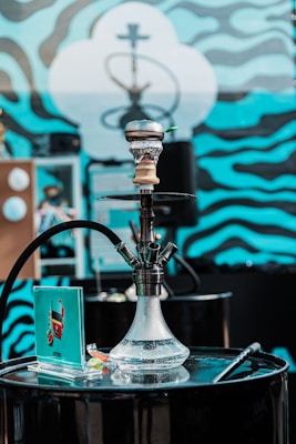 A hookah set on a black, cylindrical table with a vibrant background featuring wavy blue and black patterns. The hookah is transparent with water inside and has a hose attached. There is a small booklet or card near the base of the hookah along with a few small items.