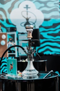 A hookah set on a black, cylindrical table with a vibrant background featuring wavy blue and black patterns. The hookah is transparent with water inside and has a hose attached. There is a small booklet or card near the base of the hookah along with a few small items.