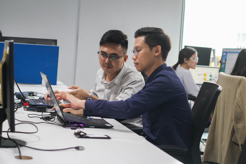 two men sitting at a desk looking at a laptop