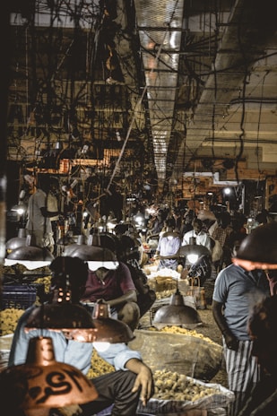 A busy market scene with vendors selling their goods under low-hanging lights. The ceiling is lined with wires and cables, and the market is bustling with people and vibrant activity.