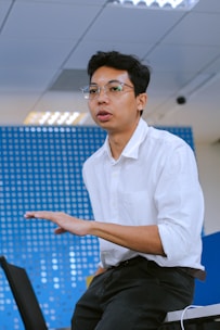 A person wearing glasses and a white shirt is sitting on a chair with a hand gesturing forward. The background is a blue wall with circular patterns and a ceiling with bright lights.