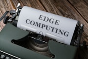 A vintage typewriter holds a sheet of paper with the words 'EDGE COMPUTING' typed in bold letters. The typewriter sits on a rustic wooden surface, giving a retro yet modern technological feel.