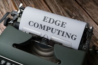 A vintage typewriter holds a sheet of paper with the words 'EDGE COMPUTING' typed in bold letters. The typewriter sits on a rustic wooden surface, giving a retro yet modern technological feel.
