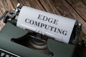 A vintage typewriter holds a sheet of paper with the words 'EDGE COMPUTING' typed in bold letters. The typewriter sits on a rustic wooden surface, giving a retro yet modern technological feel.