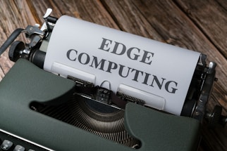 A vintage typewriter holds a sheet of paper with the words 'EDGE COMPUTING' typed in bold letters. The typewriter sits on a rustic wooden surface, giving a retro yet modern technological feel.