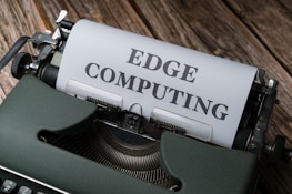 A vintage typewriter holds a sheet of paper with the words 'EDGE COMPUTING' typed in bold letters. The typewriter sits on a rustic wooden surface, giving a retro yet modern technological feel.