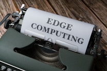 A vintage typewriter holds a sheet of paper with the words 'EDGE COMPUTING' typed in bold letters. The typewriter sits on a rustic wooden surface, giving a retro yet modern technological feel.