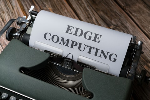 A vintage typewriter holds a sheet of paper with the words 'EDGE COMPUTING' typed in bold letters. The typewriter sits on a rustic wooden surface, giving a retro yet modern technological feel.