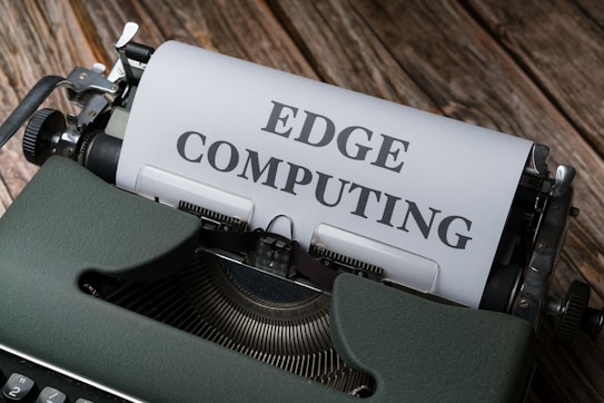 A vintage typewriter holds a sheet of paper with the words 'EDGE COMPUTING' typed in bold letters. The typewriter sits on a rustic wooden surface, giving a retro yet modern technological feel.