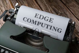 A vintage typewriter holds a sheet of paper with the words 'EDGE COMPUTING' typed in bold letters. The typewriter sits on a rustic wooden surface, giving a retro yet modern technological feel.