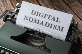 A vintage typewriter with a sheet of paper in its platen. The paper has the words 'DIGITAL NOMADISM' typed in bold, clear letters. The typewriter is placed on a rustic wooden surface, conveying a blend of nostalgia and modernity.