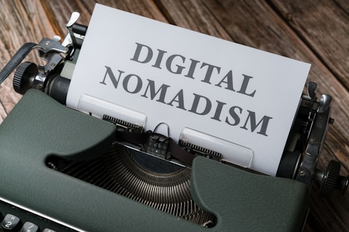 A vintage typewriter with a sheet of paper in its platen. The paper has the words 'DIGITAL NOMADISM' typed in bold, clear letters. The typewriter is placed on a rustic wooden surface, conveying a blend of nostalgia and modernity.