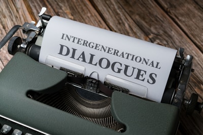 A vintage typewriter with a piece of paper inserted, displaying the text 'INTERGENERATIONAL DIALOGUES'. The typewriter is set on a rustic wooden surface, and the paper is visible above the keys.