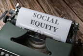A vintage typewriter with a sheet of paper that has the words 'SOCIAL EQUITY' typed in bold letters. The typewriter is placed on a rustic wooden surface.
