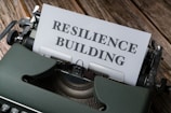 An old-fashioned typewriter holds a sheet of paper with the words 'RESILIENCE BUILDING' typed on it. The machine sits on a rustic wooden table, and the typewriter features a sleek, dark green exterior with metallic components.