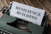 An old-fashioned typewriter holds a sheet of paper with the words 'RESILIENCE BUILDING' typed on it. The machine sits on a rustic wooden table, and the typewriter features a sleek, dark green exterior with metallic components.
