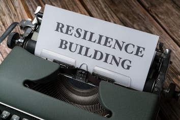 An old-fashioned typewriter holds a sheet of paper with the words 'RESILIENCE BUILDING' typed on it. The machine sits on a rustic wooden table, and the typewriter features a sleek, dark green exterior with metallic components.
