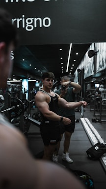 A muscular man in a black tank top poses in front of a mirror in a gym, while another person takes a photo. The gym setting includes weights and gym equipment, with a sleek and modern interior featuring dark tones and spotlights. The scene reflects in the mirror, emphasizing the symmetry of the setting.