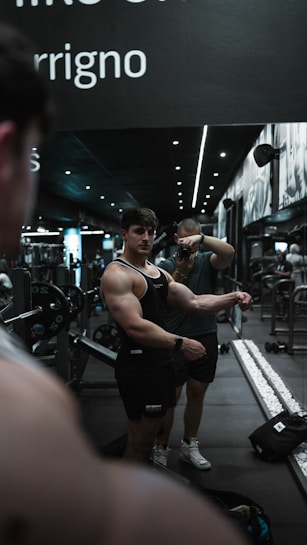 A muscular man in a black tank top poses in front of a mirror in a gym, while another person takes a photo. The gym setting includes weights and gym equipment, with a sleek and modern interior featuring dark tones and spotlights. The scene reflects in the mirror, emphasizing the symmetry of the setting.