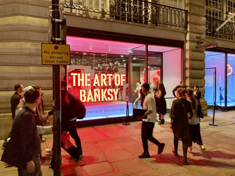 A group of people gather outside a gallery with a large illuminated display reading 'The Art of Banksy' in bold red letters. The gallery has bright red and blue lighting inside, creating a vibrant and dynamic atmosphere. The exterior architecture features large windows and ornate stonework. A sign nearby reads 'No stopping except local buses.'