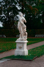A classical marble statue depicting a partially draped figure, positioned on a pedestal in a garden setting. The statue is surrounded by lush greenery and is placed along a gravel path with neatly trimmed grass. The background features tall trees and foliage displaying a mix of green and autumn tones.