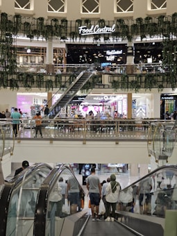 The image captures a busy indoor shopping mall setting with multiple levels. An escalator descends from the upper floor where several people can be seen carrying shopping bags. On the upper level, there is a dining area named 'Food Central' that features greenery decor and lighted signage. Numerous shops with bright displays and signage are also visible in the background.