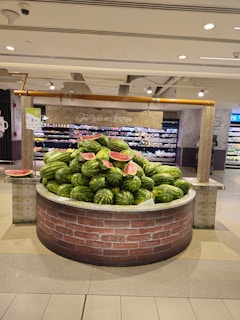 A large stack of watermelons is displayed on a circular brick structure resembling a well in a grocery store. Some watermelons have been cut open, displaying their bright red insides. The surrounding area has shelves with various products, and there are signs with price tags.