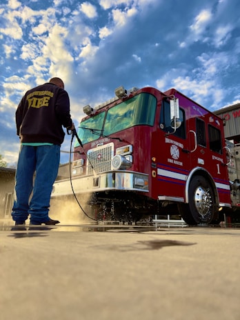 A firefighter is washing a red fire truck with a hose. The truck has prominent chrome details and various equipment attached. The sky is partially cloudy and the ground is wet, reflecting the scene above.