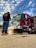 A firefighter is washing a red fire truck with a hose. The truck has prominent chrome details and various equipment attached. The sky is partially cloudy and the ground is wet, reflecting the scene above.