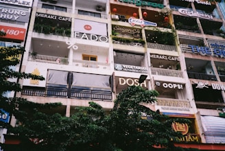 The image displays a multi-story building facade with various cafes, bars, and restaurants. The establishments have large signs with different names and logos. There are balconies with railings, some decorated with plants and red lanterns. The lower section of the building is partially obscured by leafy trees.