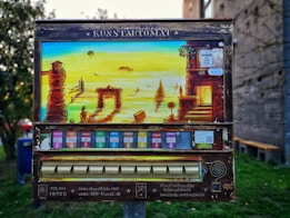 An art vending machine, labeled 'Kunstautomat,' features a vibrant painting of a surreal landscape with ancient ruins, figures, and a body of water. Below the painting, colorful slots for art packets are visible.