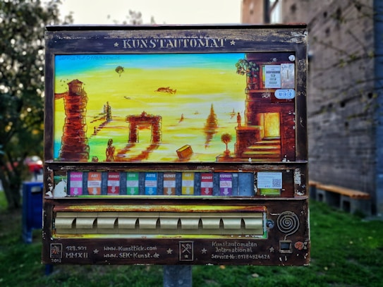 An art vending machine, labeled 'Kunstautomat,' features a vibrant painting of a surreal landscape with ancient ruins, figures, and a body of water. Below the painting, colorful slots for art packets are visible.