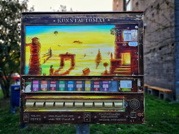 An art vending machine, labeled 'Kunstautomat,' features a vibrant painting of a surreal landscape with ancient ruins, figures, and a body of water. Below the painting, colorful slots for art packets are visible.