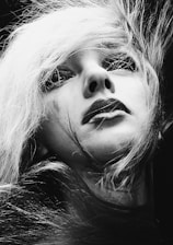 A black and white portrait features a close-up of a person with striking makeup and long, flowing hair. The expression appears to be contemplative or introspective, with the person's gaze directed slightly upward. The lighting highlights the contrast between the light strands of hair and the shadows on the face, creating a dramatic effect.
