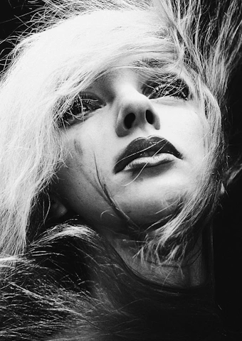 A black and white portrait features a close-up of a person with striking makeup and long, flowing hair. The expression appears to be contemplative or introspective, with the person's gaze directed slightly upward. The lighting highlights the contrast between the light strands of hair and the shadows on the face, creating a dramatic effect.
