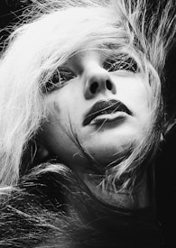 A black and white portrait features a close-up of a person with striking makeup and long, flowing hair. The expression appears to be contemplative or introspective, with the person's gaze directed slightly upward. The lighting highlights the contrast between the light strands of hair and the shadows on the face, creating a dramatic effect.