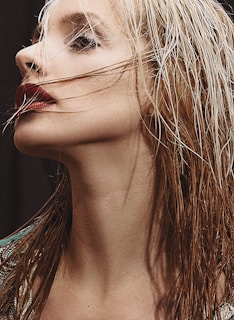 A close-up profile of a person with light-colored wet hair. The person's makeup is bold, featuring dark red lipstick and prominent eye makeup. Strands of hair partially cover the face, adding a dramatic touch.