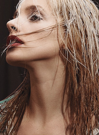 A close-up profile of a person with light-colored wet hair. The person's makeup is bold, featuring dark red lipstick and prominent eye makeup. Strands of hair partially cover the face, adding a dramatic touch.