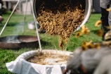 A person is pouring a mixture of herbs or plant material from a container through a filter setup over a basin. The scene is set outdoors on a green lawn with various equipment in the background, including a metal basin and a tripod structure.