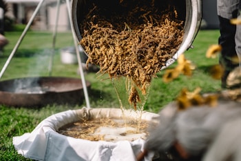 A person is pouring a mixture of herbs or plant material from a container through a filter setup over a basin. The scene is set outdoors on a green lawn with various equipment in the background, including a metal basin and a tripod structure.