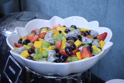 A large white ceramic bowl contains a fresh fruit salad, featuring a mix of colorful fruits such as strawberries, grape varieties, dragon fruit, mango, and green grapes. The fruit pieces are cut into bite-sized portions, arranged in a visually appealing manner.