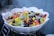 A large white ceramic bowl contains a fresh fruit salad, featuring a mix of colorful fruits such as strawberries, grape varieties, dragon fruit, mango, and green grapes. The fruit pieces are cut into bite-sized portions, arranged in a visually appealing manner.